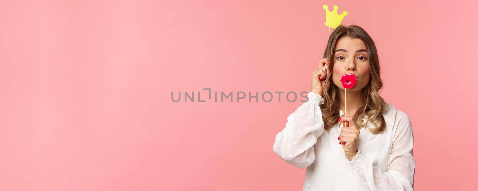 Spring, happiness and celebration concept. Close-up portrait of funny and cute, silly blond girl in white dress, holding big lips mask and crown, partying and having fun, pink background by Benzoix