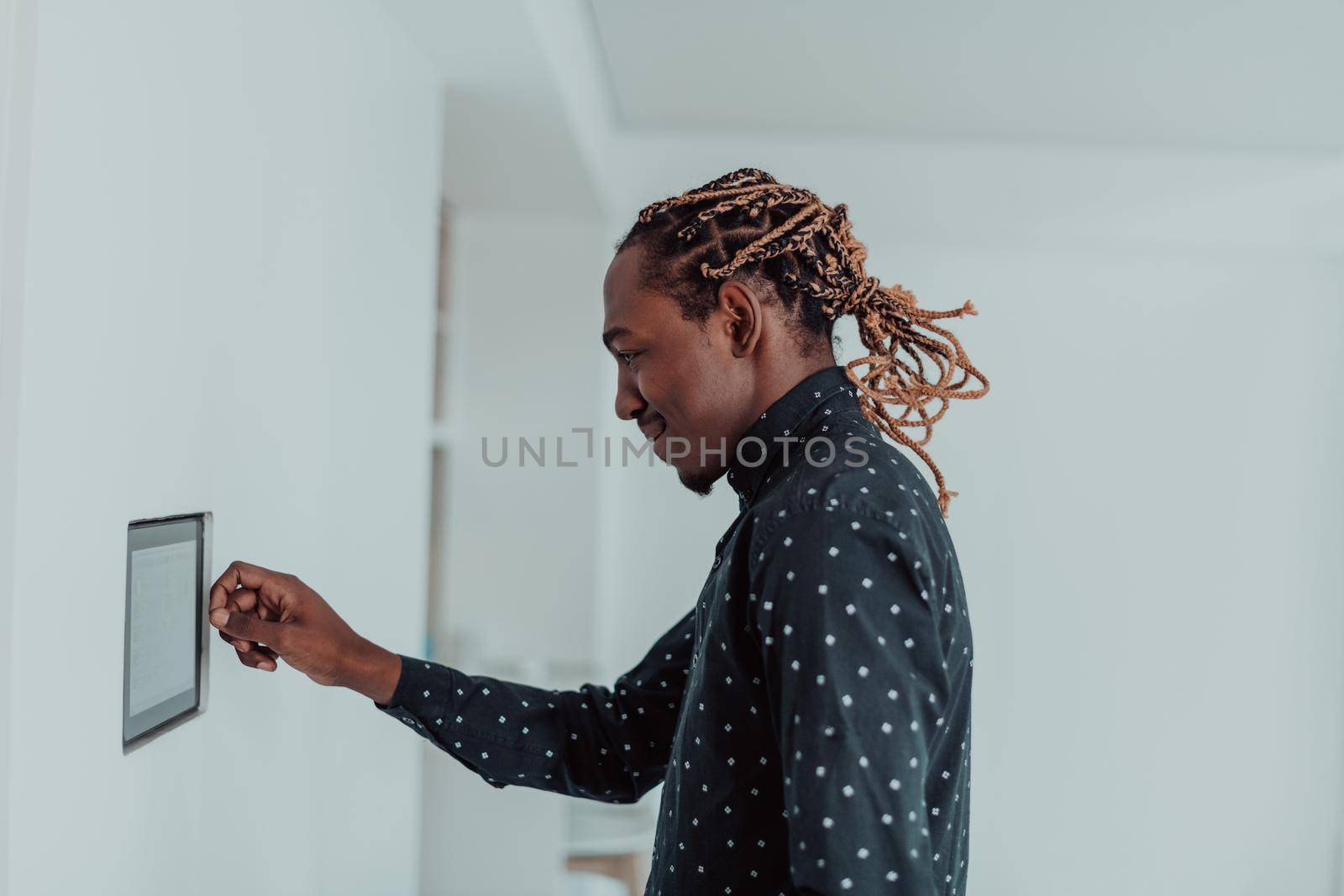 Smiling African American man using modern smart home system, controller on wall, positive young man switching temperature on thermostat or activating security alarm in apartment. High quality photo