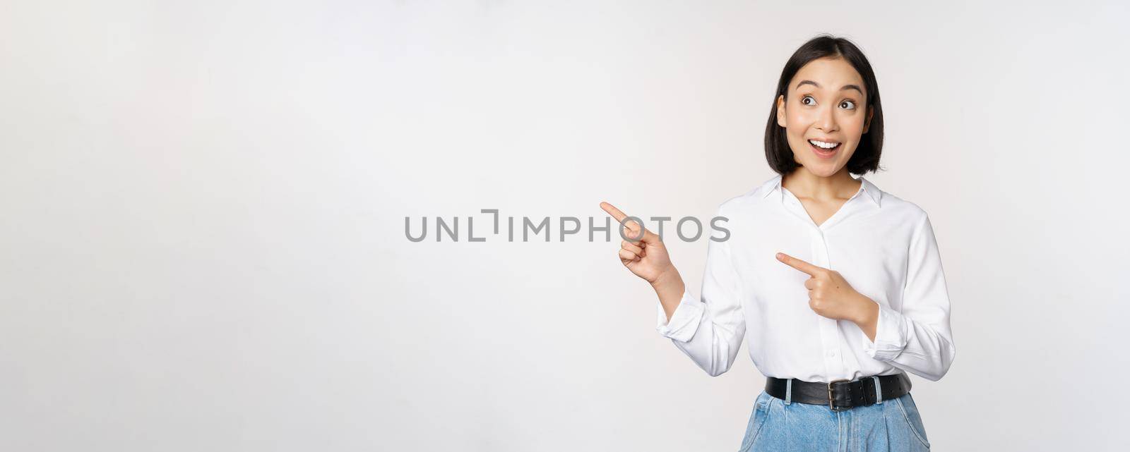 Enthusiastic korean girl pointing fingers left, female student pointing and looking left with happy smile, showing company logo or banner, white background.