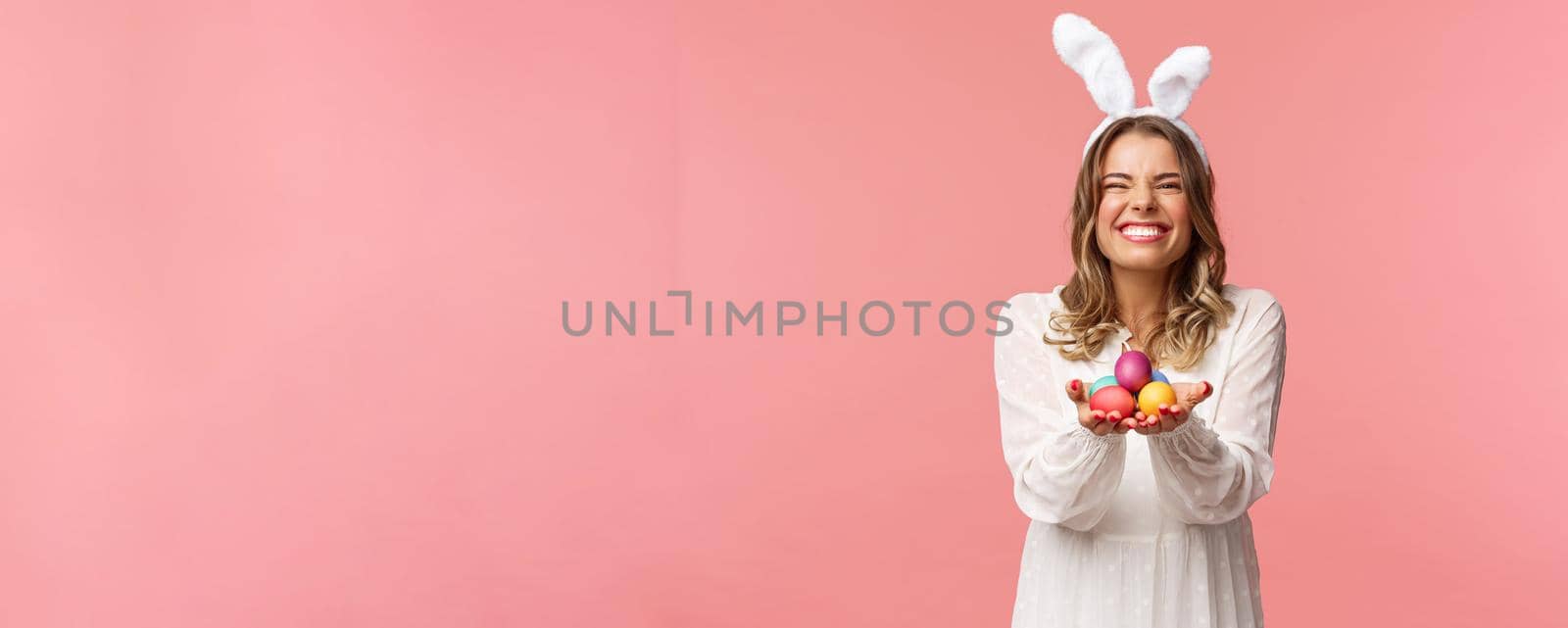 Holidays, spring and party concept. Portrait of excited charming blond woman with lovely pleased smile, holding painted eggs, wear Easter day outfit, rabbit ears and white dress, pink background.