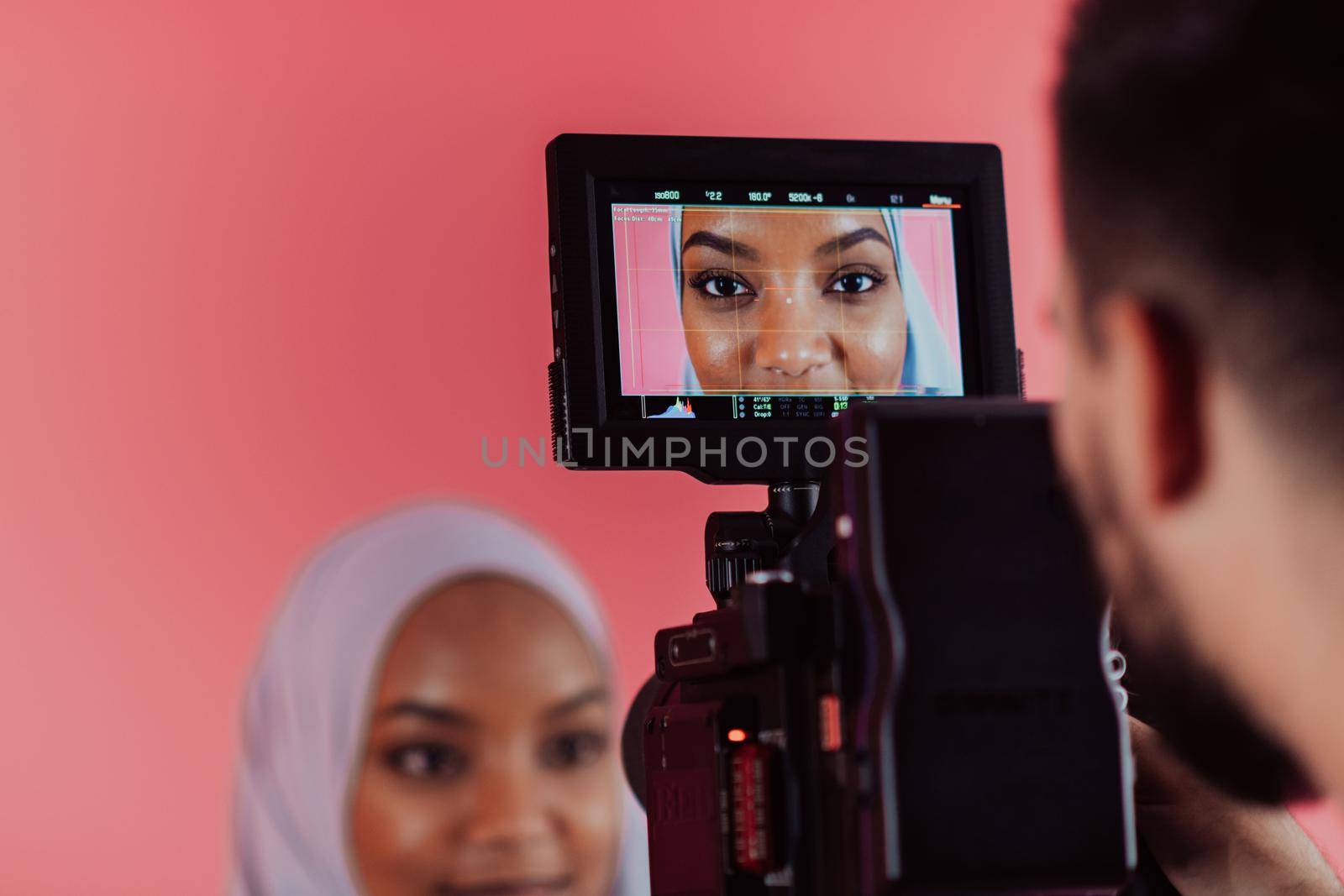 Videographer in digital studio recording video on professional camera by shooting female Muslim woman wearing hijab scarf plastic pink background. by dotshock