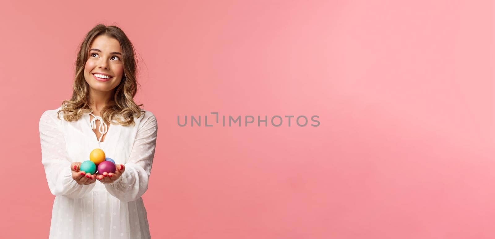 Holidays, spring and party concept. Portrait of dreamy good-looking blond girl in white dress, holding painted eggs, celebrating Easter, orthodox holy day, smiling and looking away.