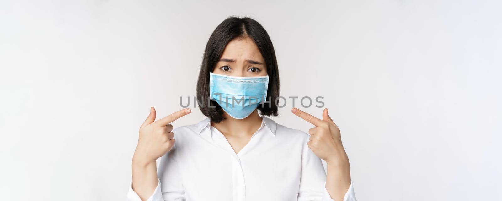 Close up of sad asian girl in medical face mask pointing at her head and looking upset, standing over white background.