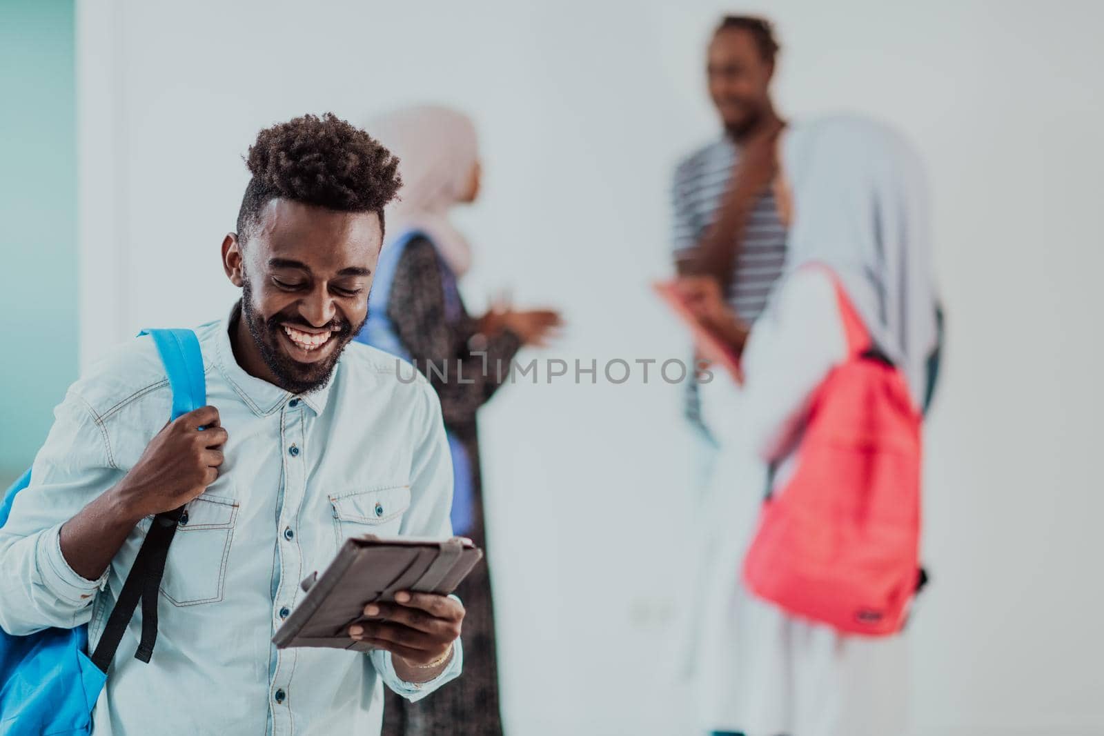 University lifestyle handsome young African student man holding a tablet computer and smiling while standing against university with his friends have a team meeting in the background. High-quality photo. High-quality photo