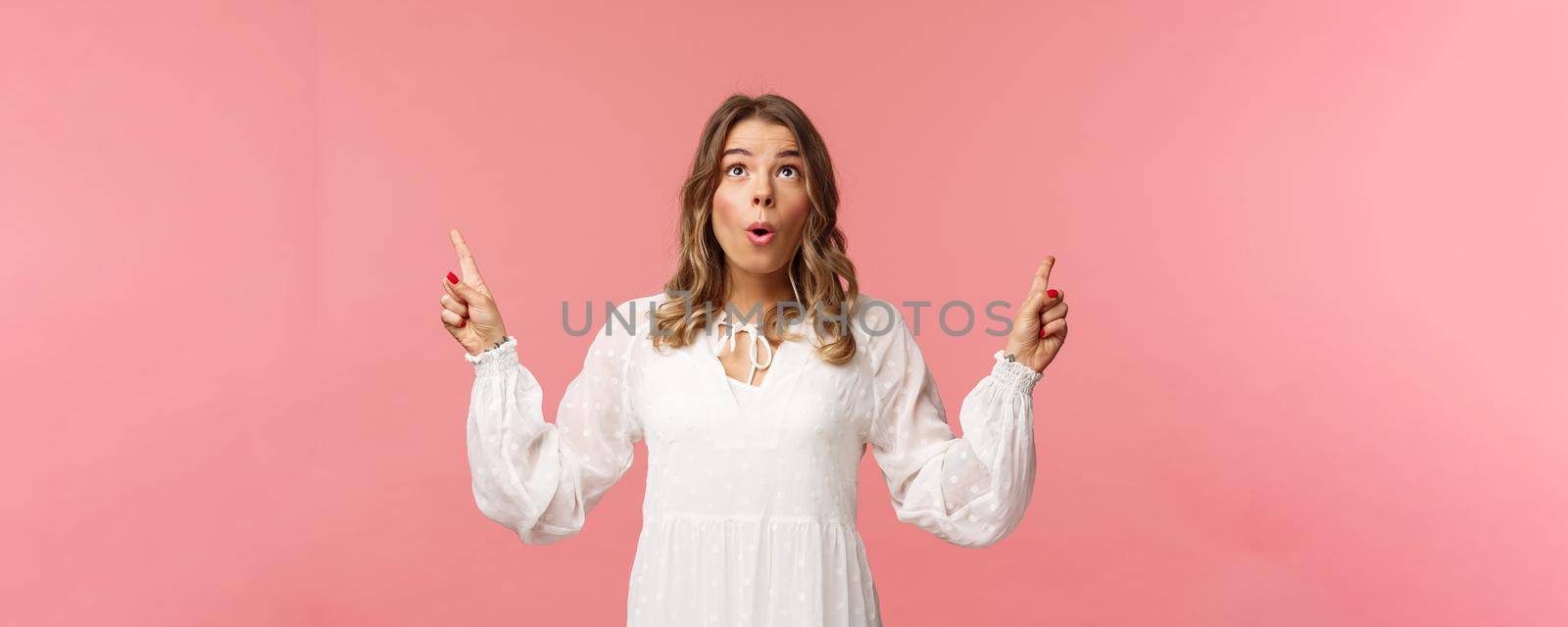 Excited beautiful caucasian blond woman in white dress, fold lips in excitement, looking pointing fingers up at something stunning and breathtaking, enjoying view, pink background by Benzoix