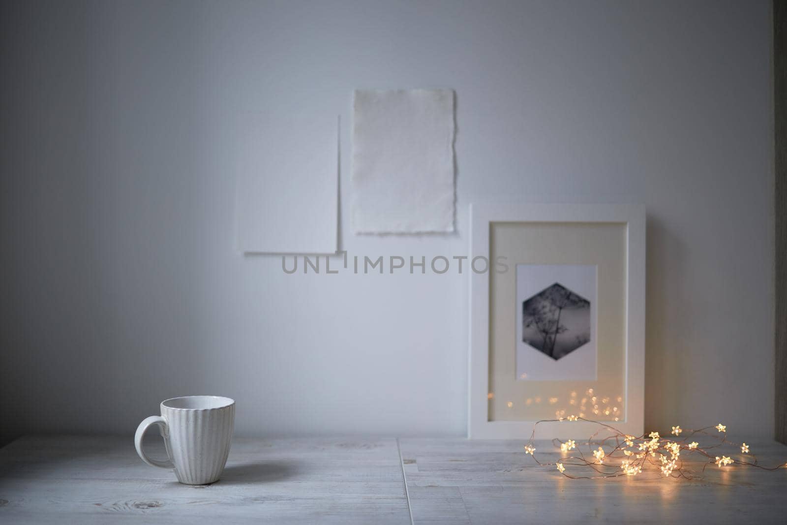 Scandinavian style. Interior Design. A white cup, cozy lights of a luminous garland, a frame for a photo are on the table. Two blank sheets of paper are attached to the wall. Empty space for text