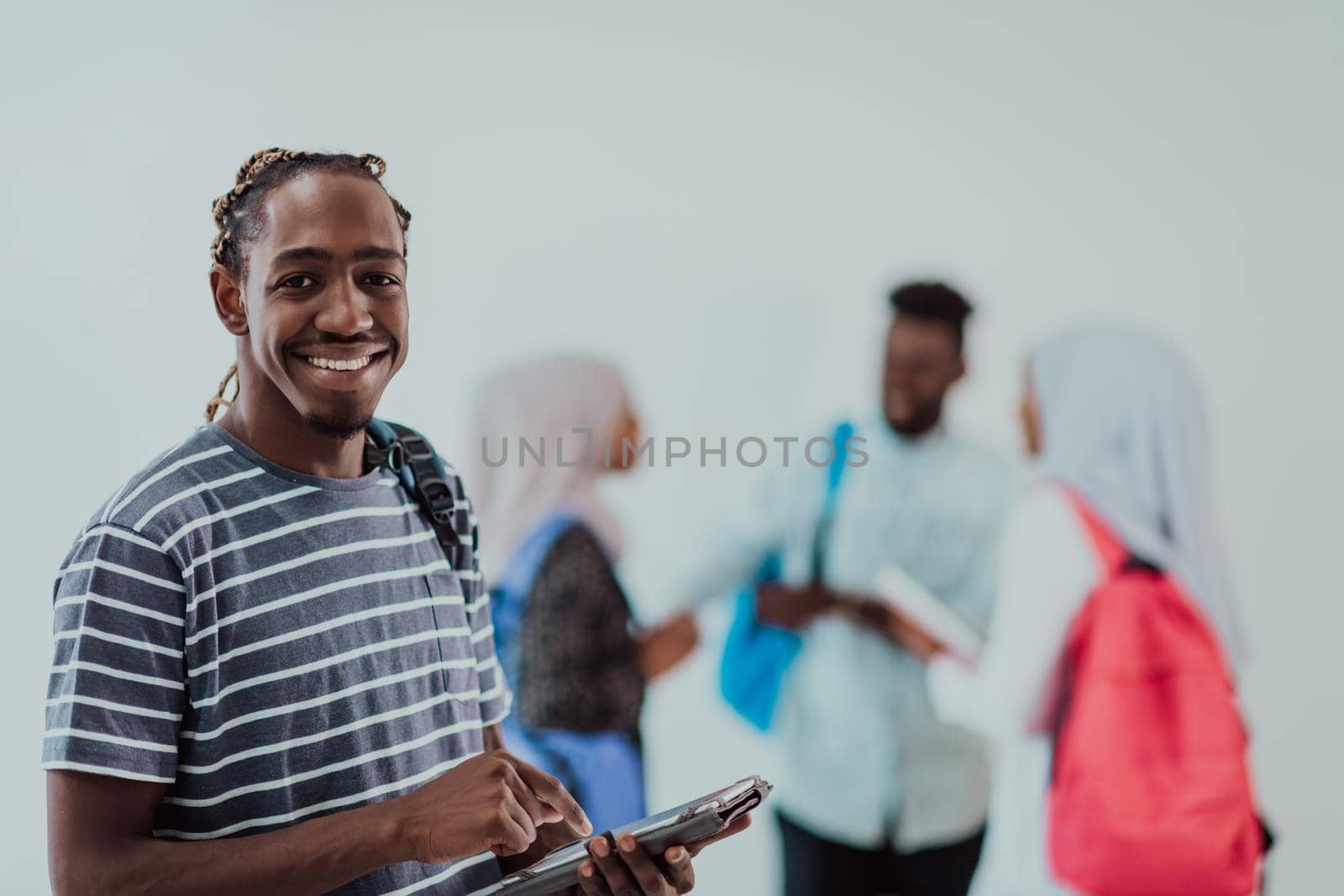 University lifestyle handsome young African student man holding a tablet computer and smiling while standing against university with his friends have a team meeting in the background. High-quality photo. High-quality photo