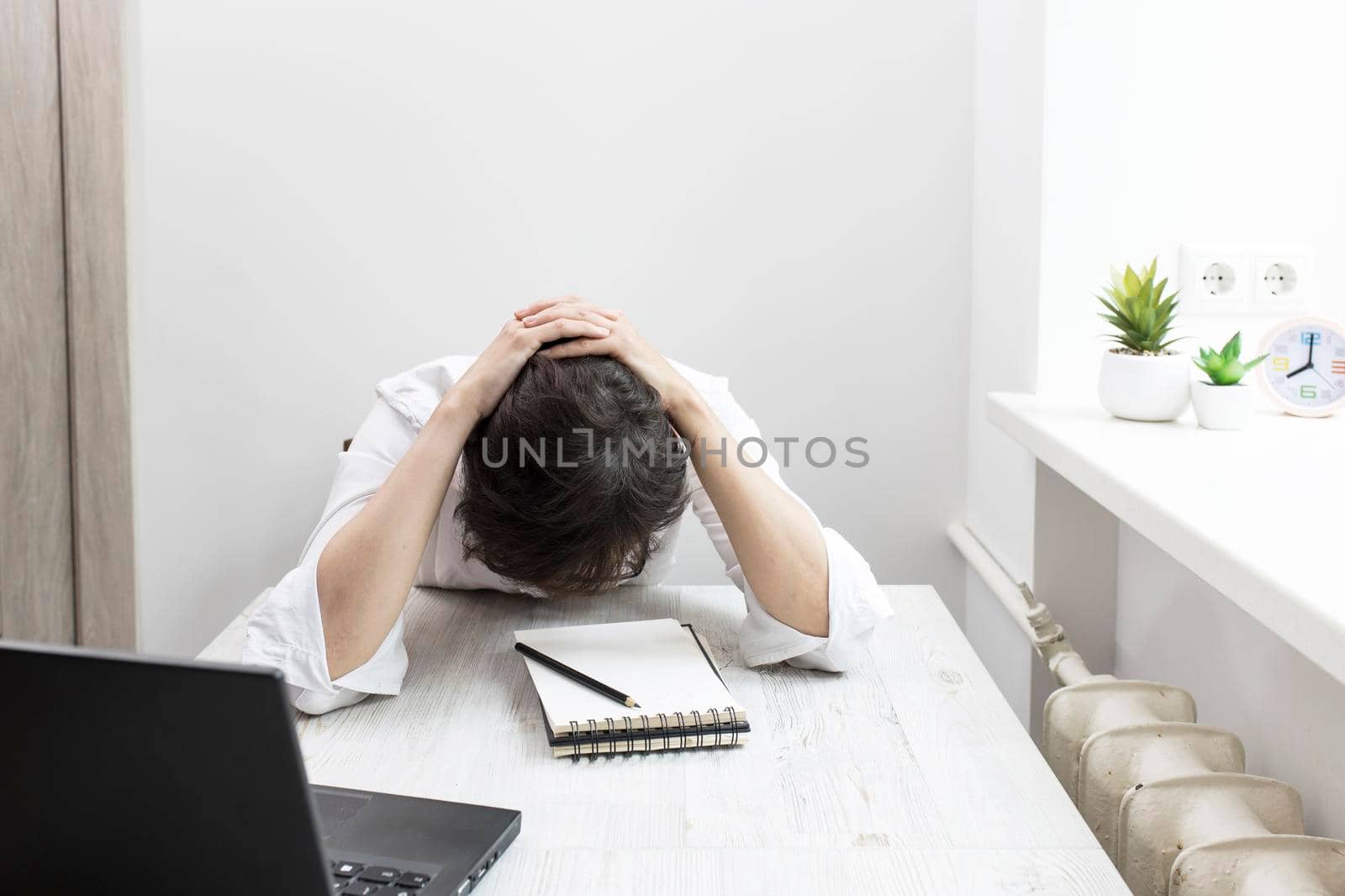 Middle-aged woman with short-haired brunette in glasses works at a laptop near window. The office worker put her head in her hands, bent over the table, the deadline has failed.