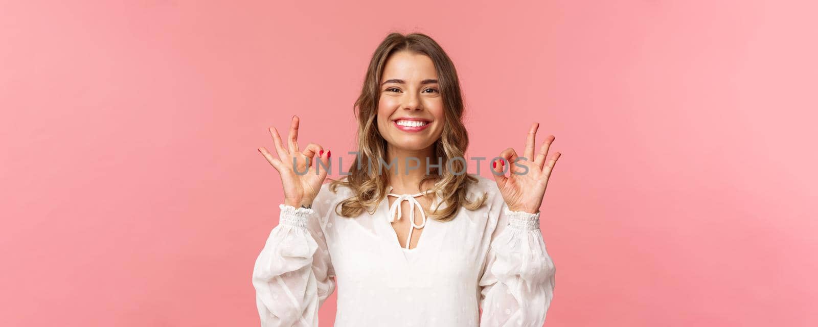 Close-up portrait of assertive, confident beautiful girl with blond shor curly hair in white dress, showing okay signs as assure no problem, smiling nod approval, recommend and guarantee quality.