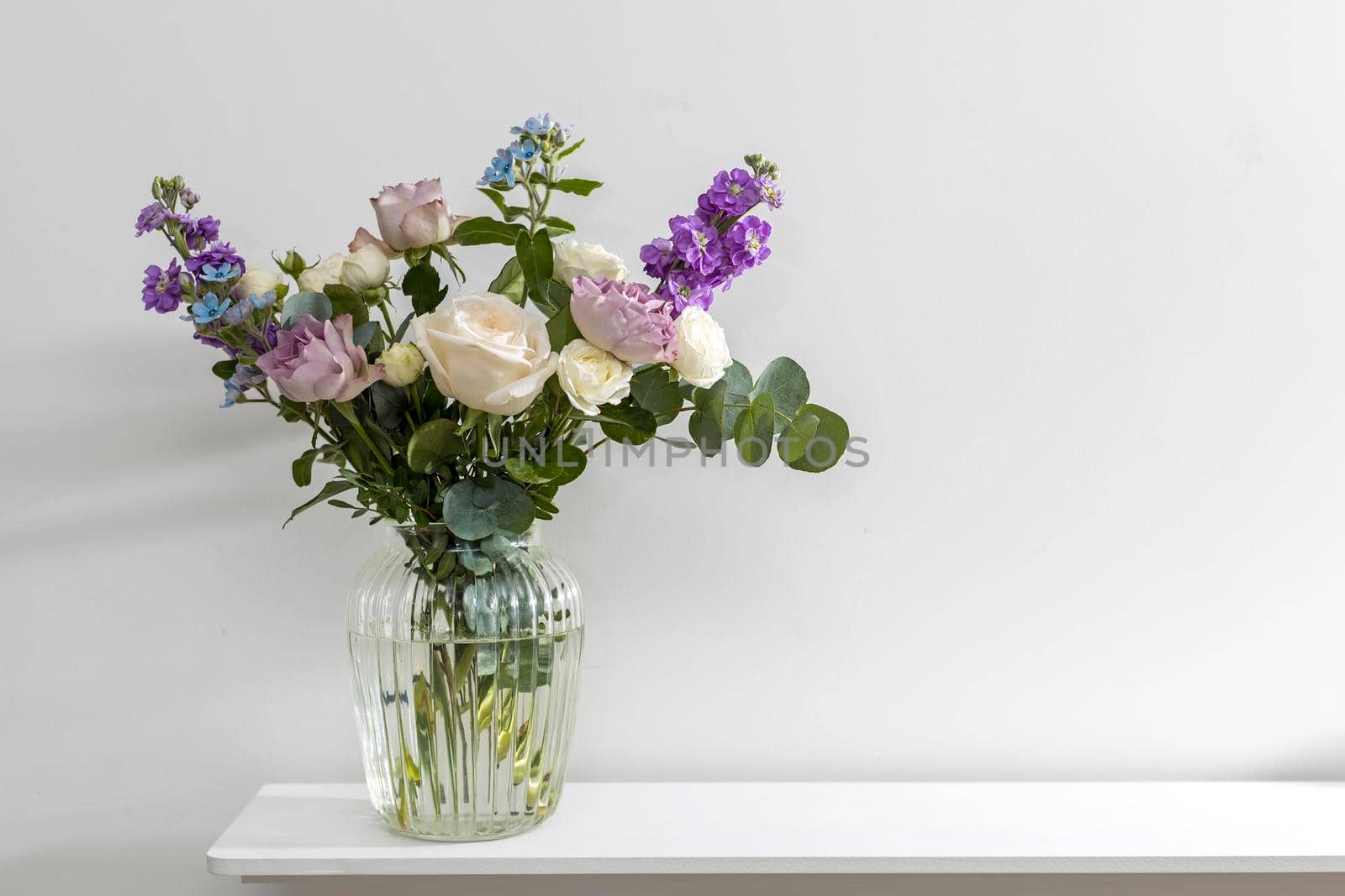 Bouquet of hackelia velutina, purple and white roses, small tea roses, matthiola incana, blue iris in glass vase is on the white coffee table . by elenarostunova