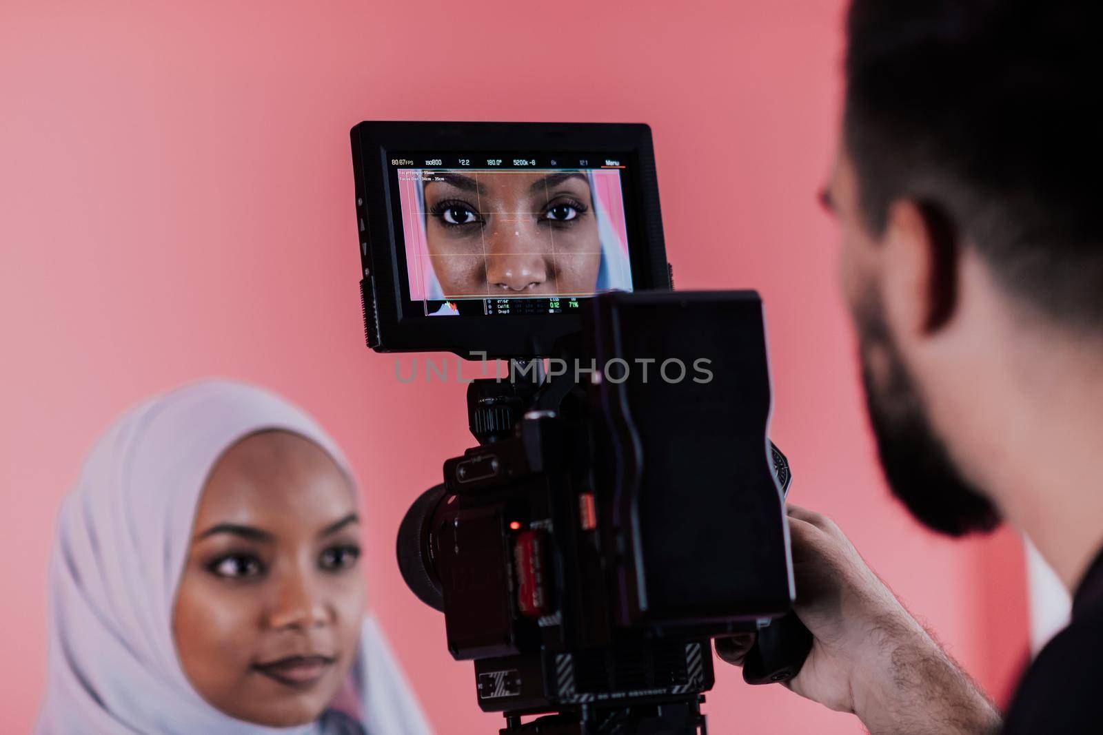 Videographer in digital studio recording video on professional camera by shooting female Muslim woman wearing hijab scarf plastic pink background. High-quality photo