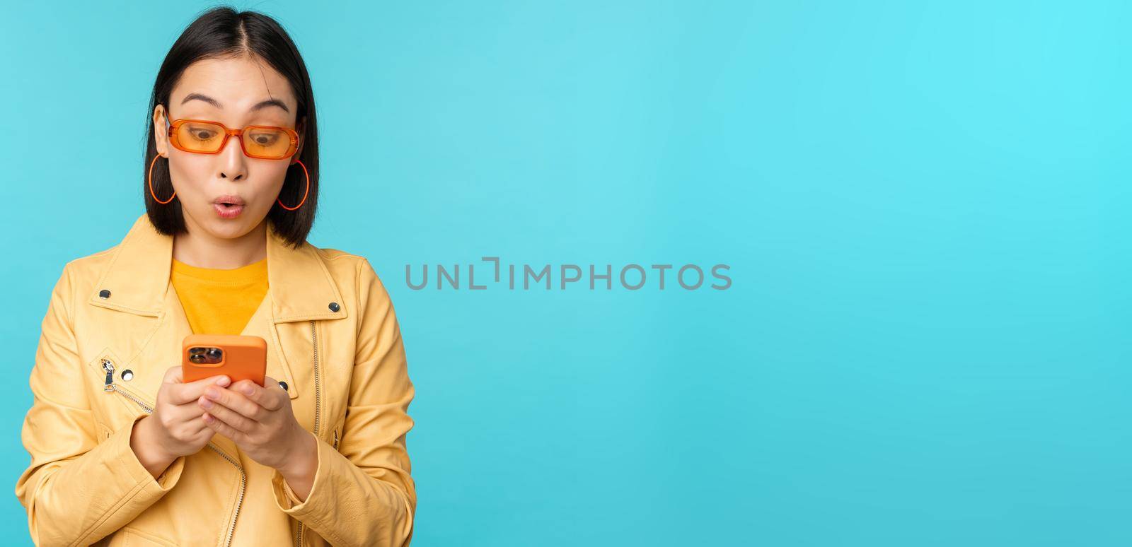 Portrait of asian girl in sunglasses, looking at smartphone screen with impressed, amazed face expression, wow face, standing over blue background by Benzoix
