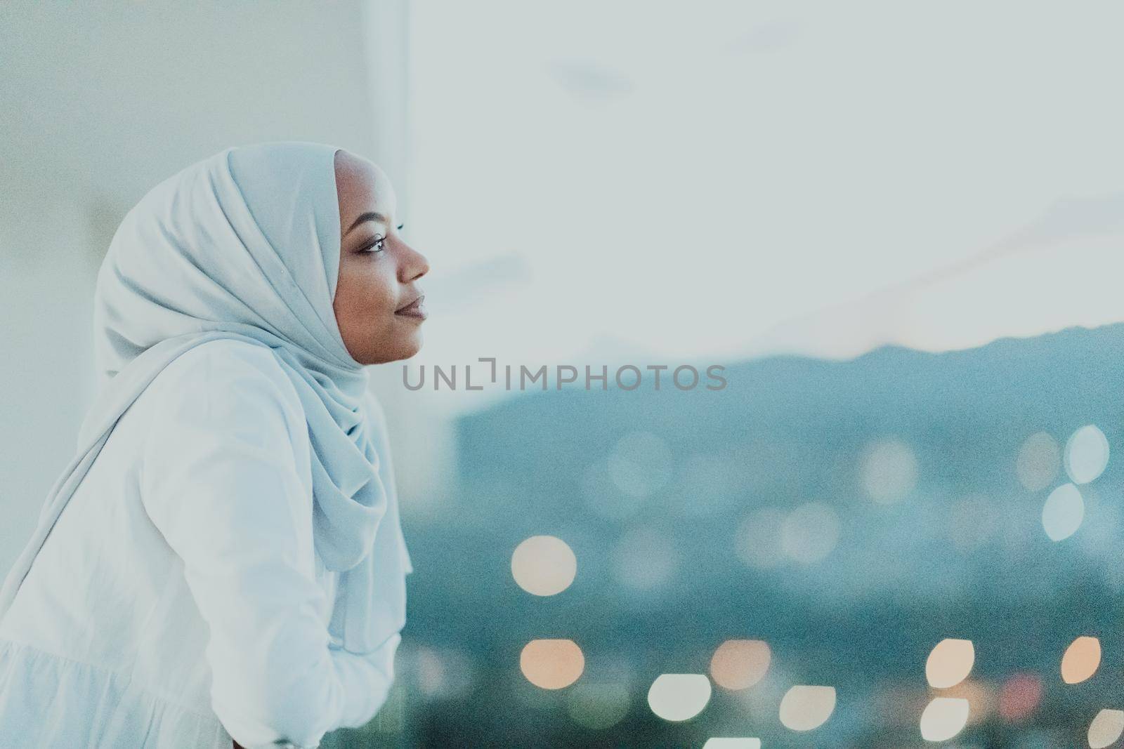 African Muslim woman in the night on a balcony smiling at the camera with city bokeh lights in the background. High-quality photo