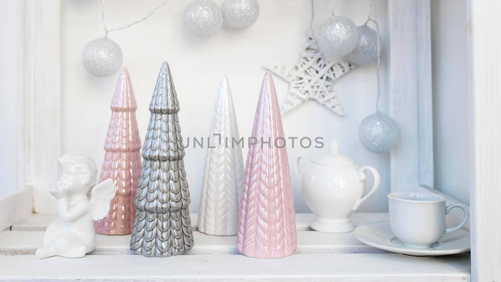 Ceramic figurines of pastel-colored Christmas trees, a white faience angel, a garland of balls made of threads decorate the room before Christmas by elenarostunova