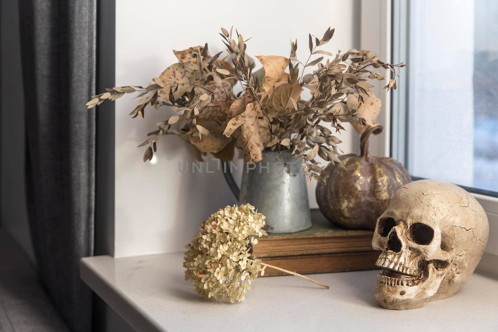Bouquet of dried autumn leaves in a zinc jug, plastic skull, artificial gilded pumpkin, vintage books decorate the window for Halloween by elenarostunova
