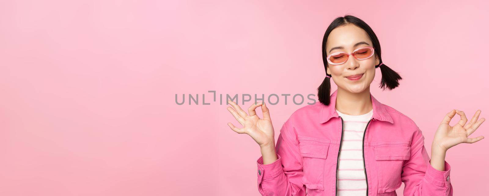 Mindfulness and wellness concept. Smiling korean girl in stylish outfit meditating, listening mantra, holds hands in zen, peace pose, practice yoga, standing over pink background.