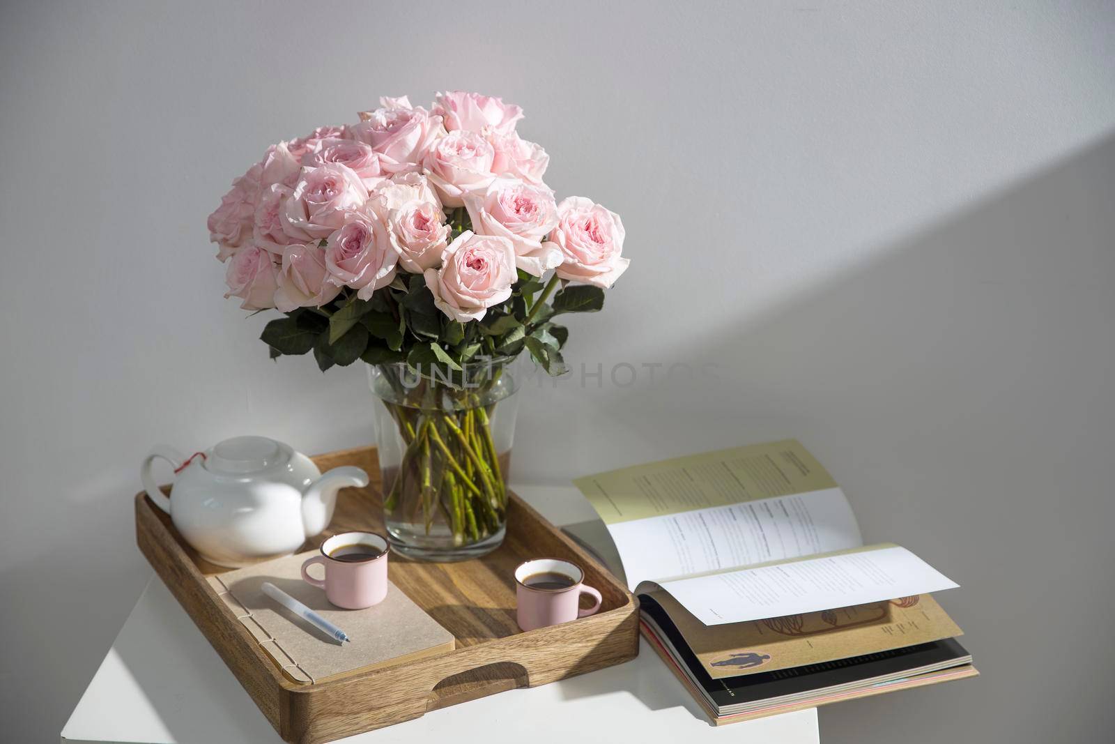 Bouquet of pink roses in a glass vase with a white teapot and two cappuccino cups on a tray on a coffee table among books by elenarostunova