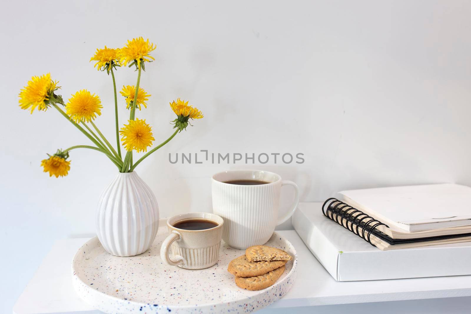 A bouquet of dandelions in a white fluted vase and two cups of different sizes with coffee on a white table. Breakfast. Copy space