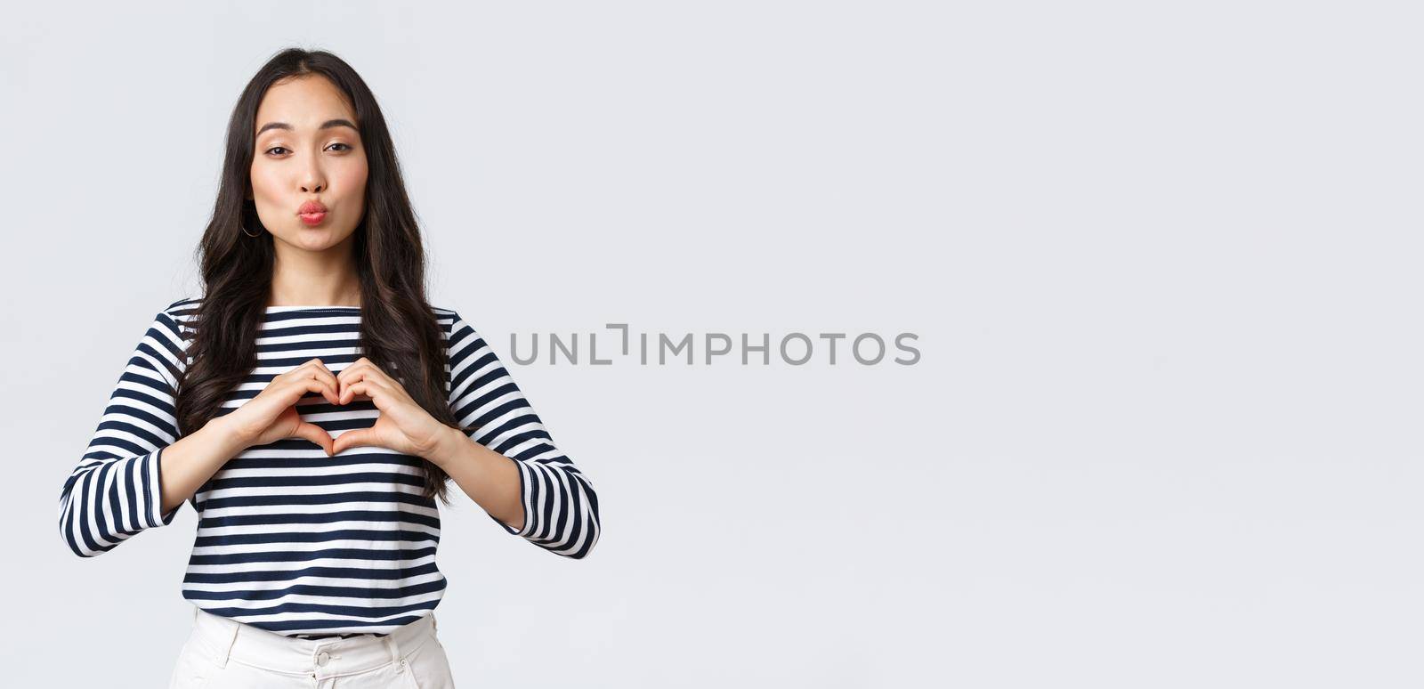 Lifestyle, people emotions and casual concept. Lovely smiling adorable asian woman showing heart sign and smiling, express sympathy or care, sending air kiss at camera, standing white background.