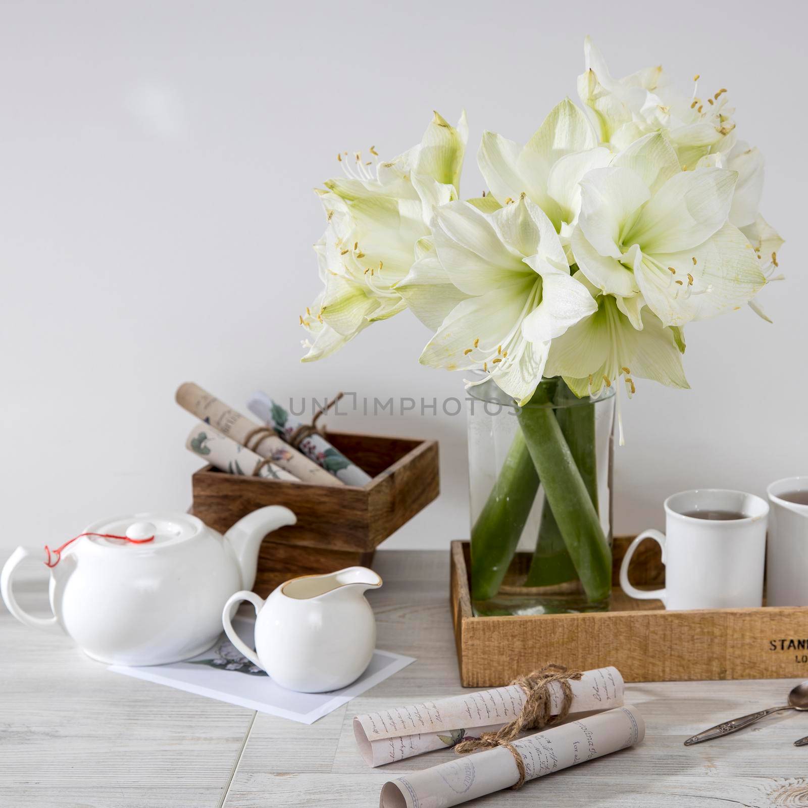 A bouquet of white lily in a glass vase on a table with two tall cups of coffee, a teapot, spoons. Copy space. Sqaure frame