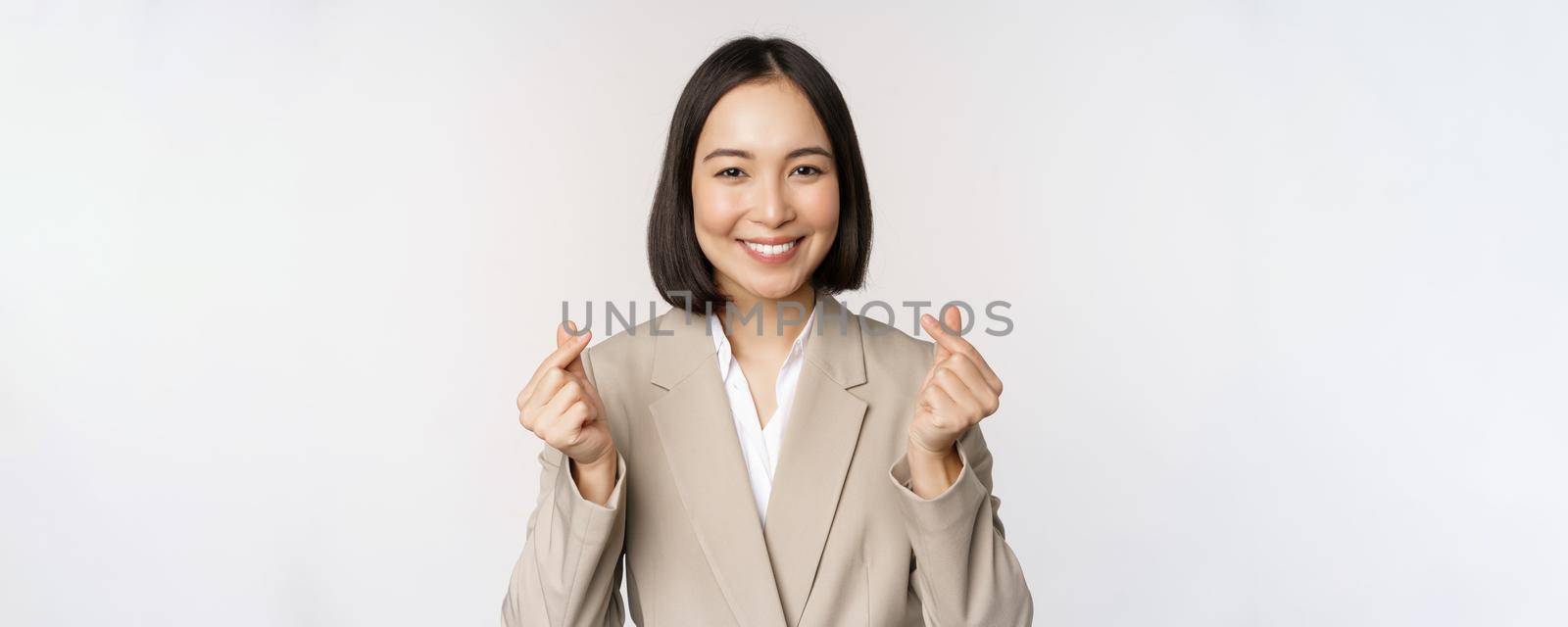 Cheerful asian saleswoman, smiling and showing finger hearts sign, standing in suit over white background.