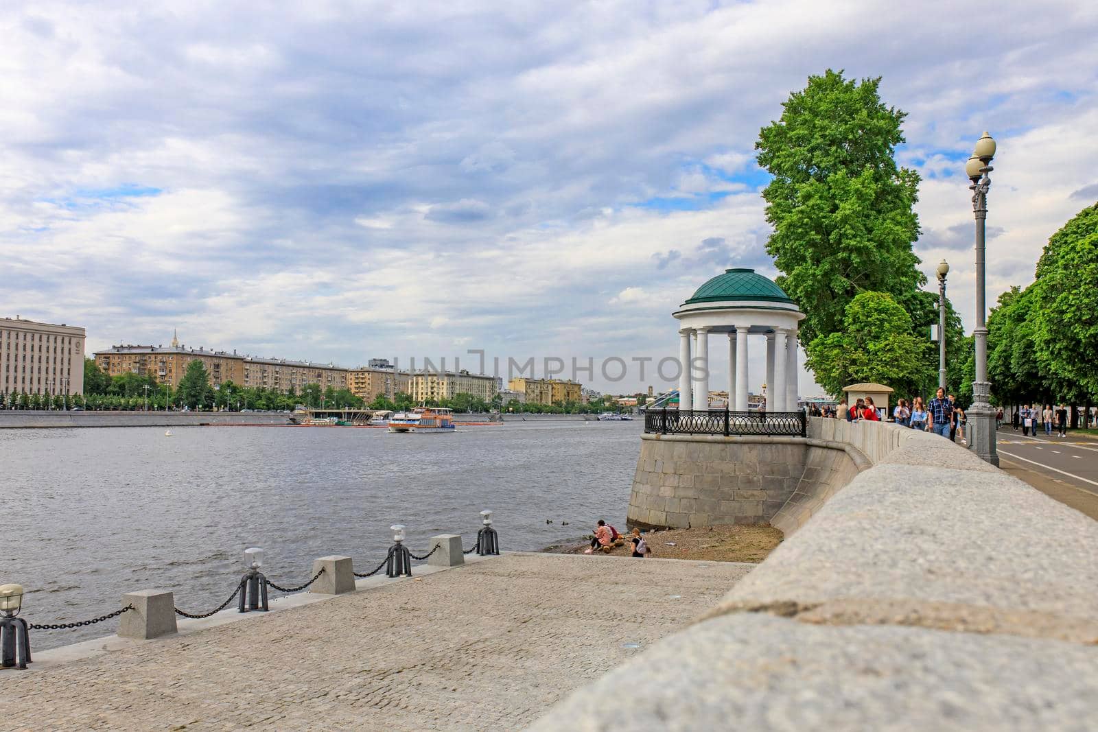 Moscow, Russia - May 15, 2021: View of Pushkin embankment. People sits on log by the river. ship sails under the bridge. Mother and child stand by the steps. Gorky Central Park of Culture and Leisure