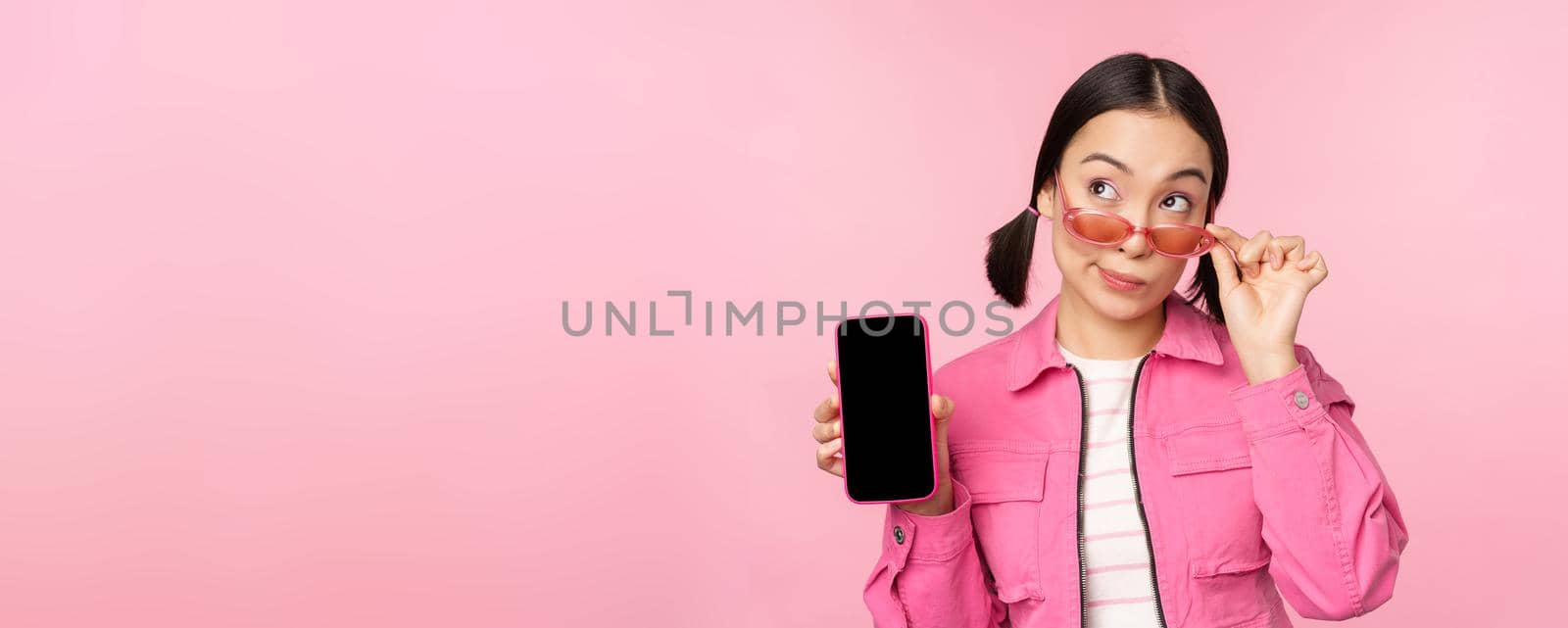 Stylish korean girl, young woman in sunglasses showing smartphone screen, mobile phone app interface or website, standing over pink background by Benzoix