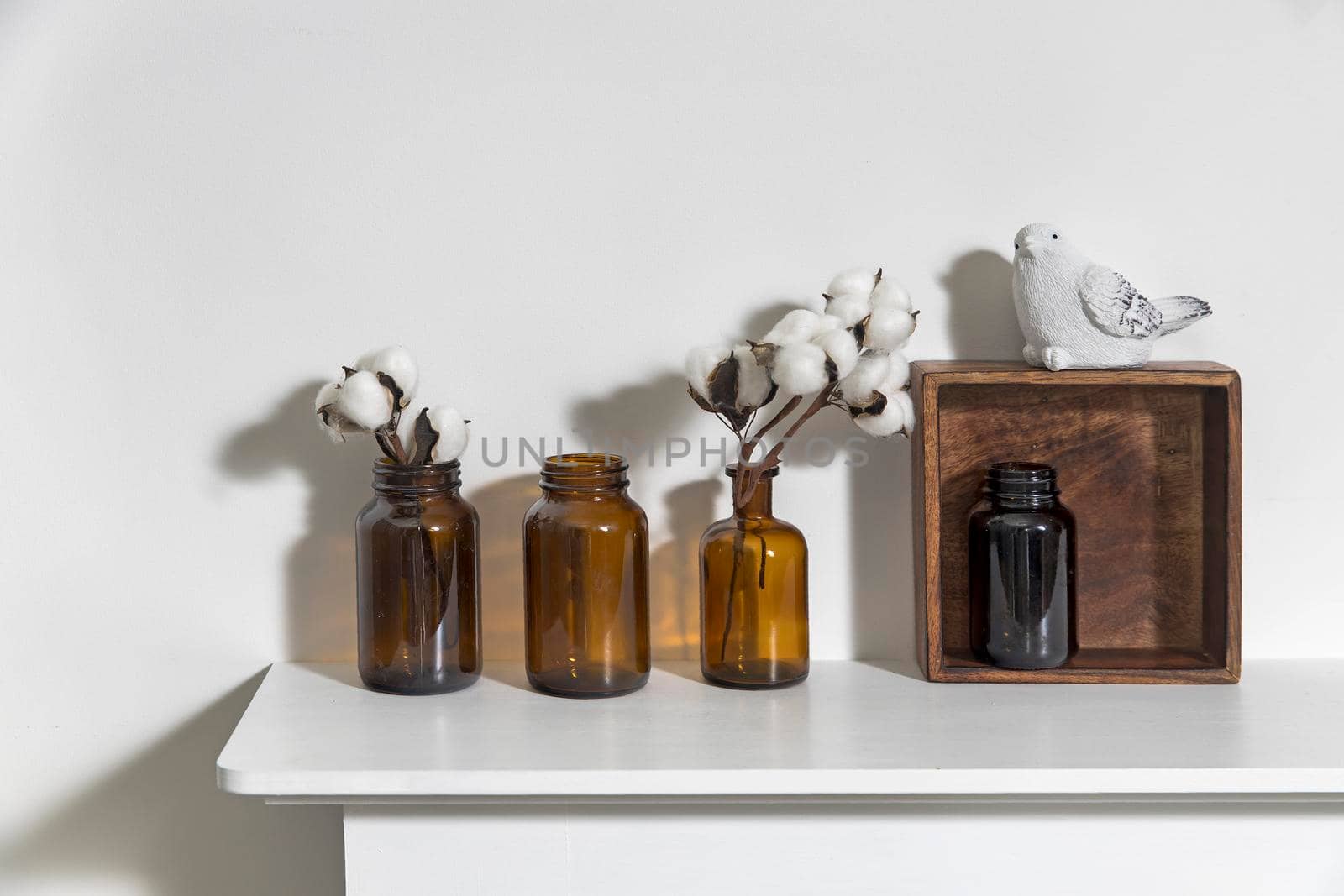 A row of brown small pharmacy bottles instead of vases with branches of blossoming cotton and wooden boxes on a white surface of the fireplace by elenarostunova
