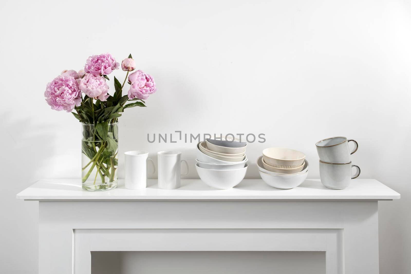 Bouquet of pink peonies, a stack of cups, plates, bowls, white chest of drawers in the interior of the kitchen by elenarostunova