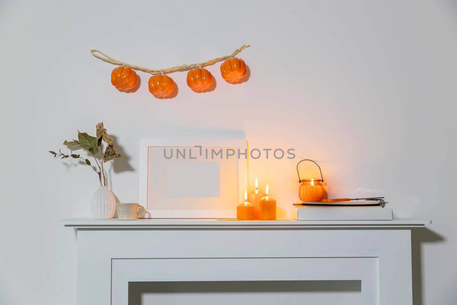 Preparing your home for Halloween. A garland of pumpkins on the wall above fake dresser panel. Frame with the inscription, orange candles and lantern. Corrugated vase with dried eucalyptus branch.