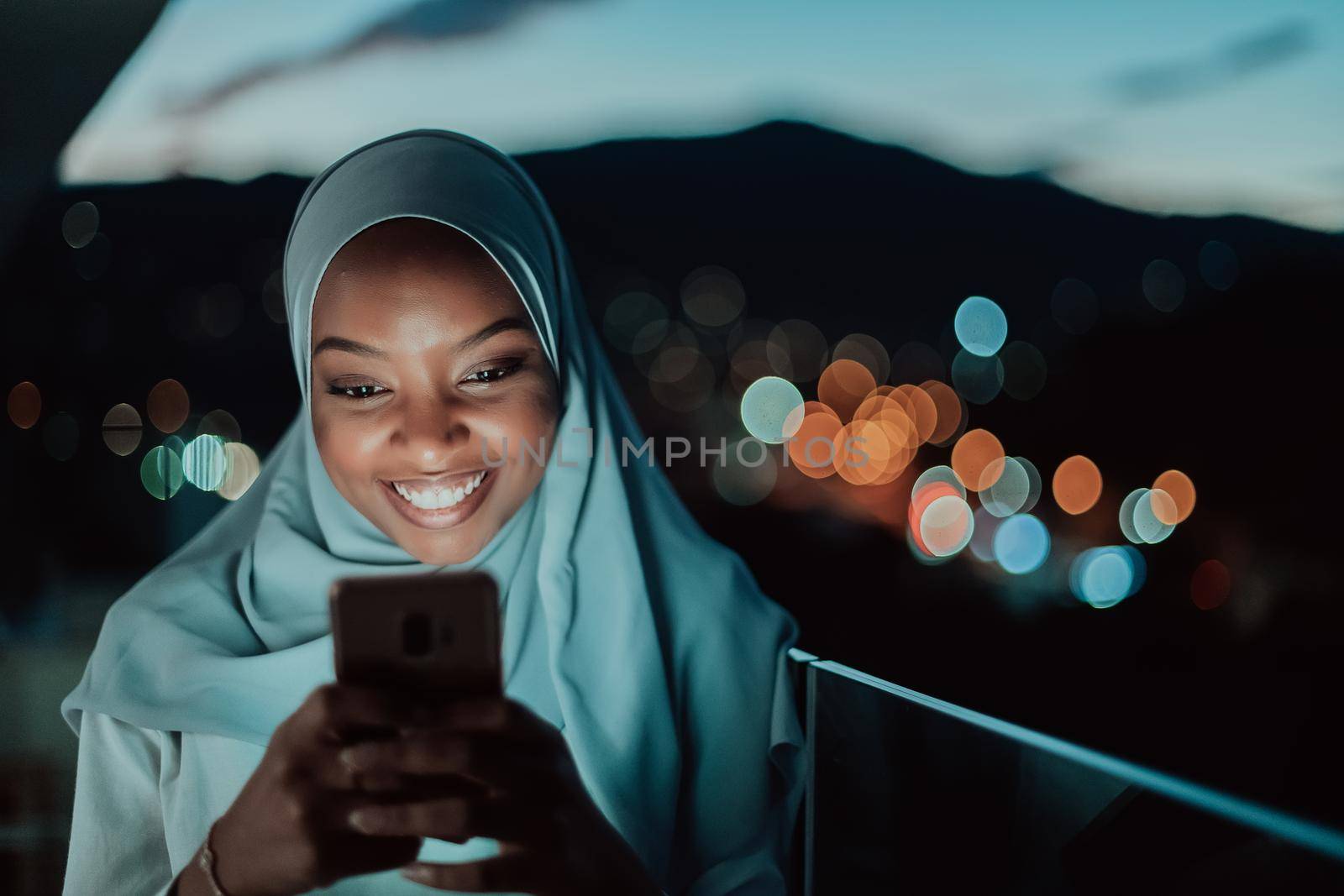 Young Muslim woman wearing scarf veil on urban city street at night texting on a smartphone with bokeh city light in the background. High-quality photo