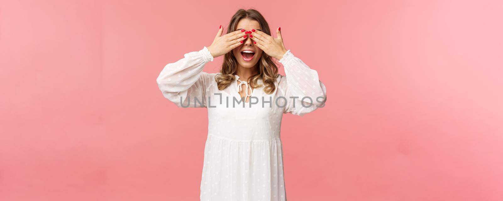 Excited cute blond girl in white spring dress waiting for gifts at surprise party, cover eyes blindfolded standing pink background, open mouth in amazement and anticipation.