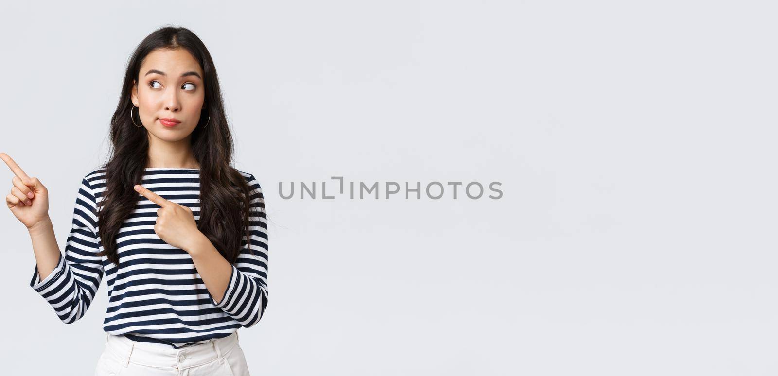 Lifestyle, beauty and fashion, people emotions concept. Curious and interested cute asian girl peeking and pointing fingers left at new promo banner with special discount in shop.