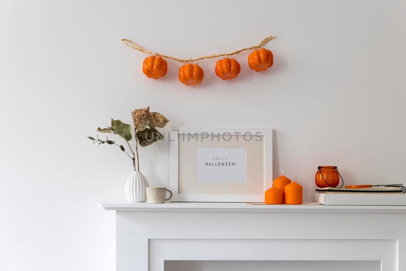 Preparing your home for Halloween. A garland of pumpkins on the wall above fake dresser panel. Frame with the inscription, orange candles and lantern. Corrugated vase with dried eucalyptus branch.