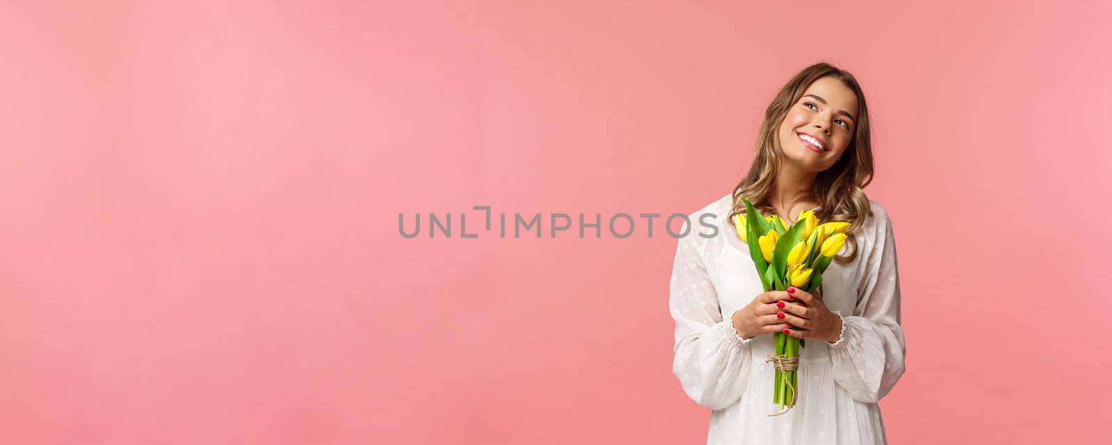 Holidays, beauty and spring concept. Portrait of dreamy, happy blond girl feeling romantic daydreaming about her girlfriend, holding yellow tulips, wear white dress, standing pink background by Benzoix