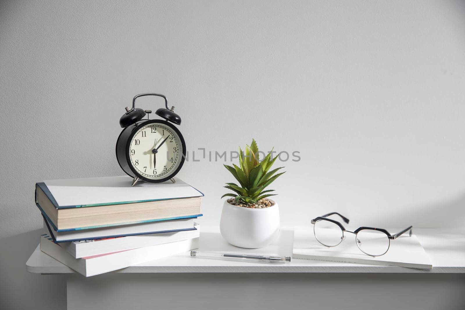 A stack of books, an artificial succulent plant in a ceramic pot, glasses, a metal basket with notebooks, a pen are on a white table surface. Concept - office at home. Online training.