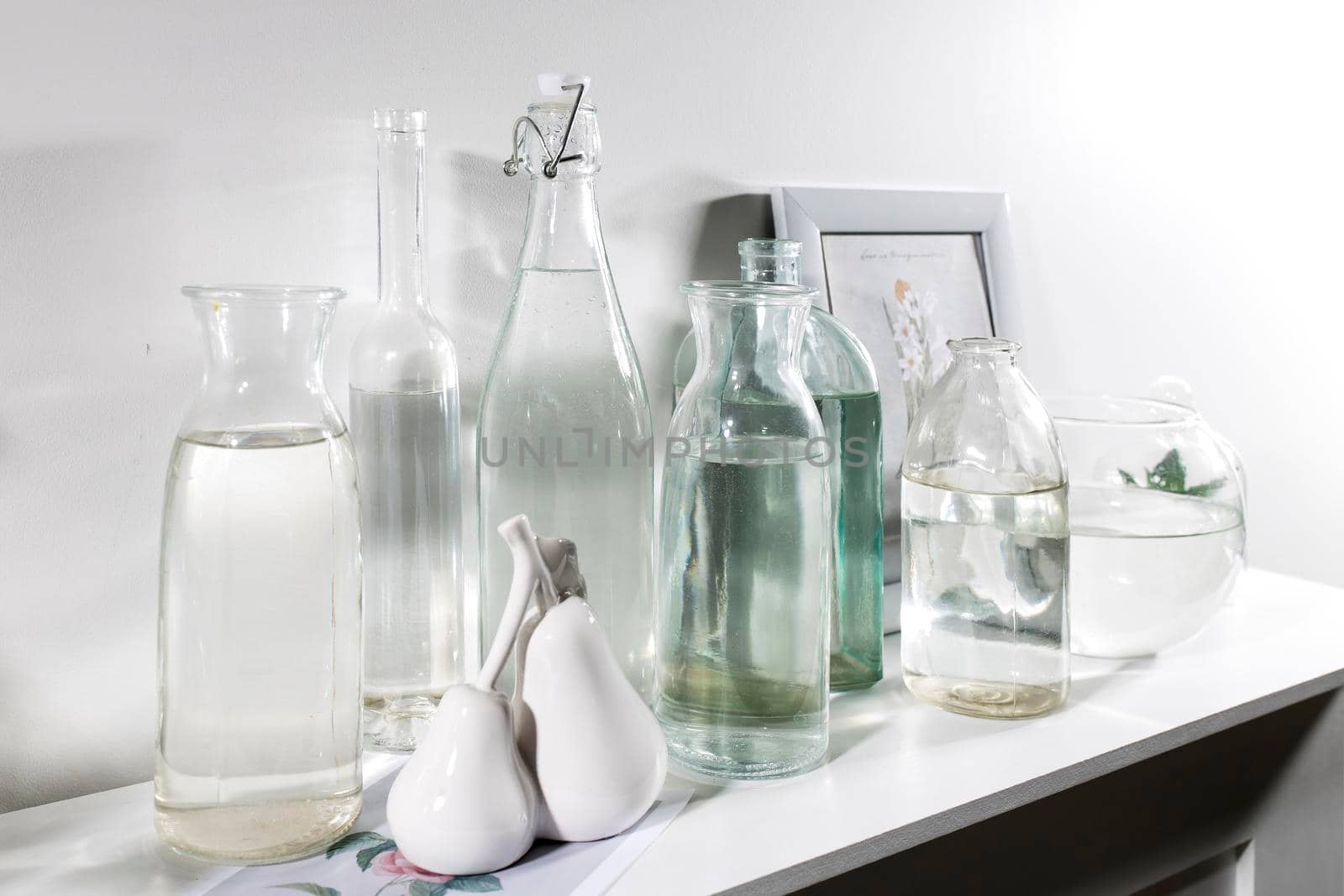 A set of bottles of different shapes with water with a photo frame is on the chest of drawers by elenarostunova