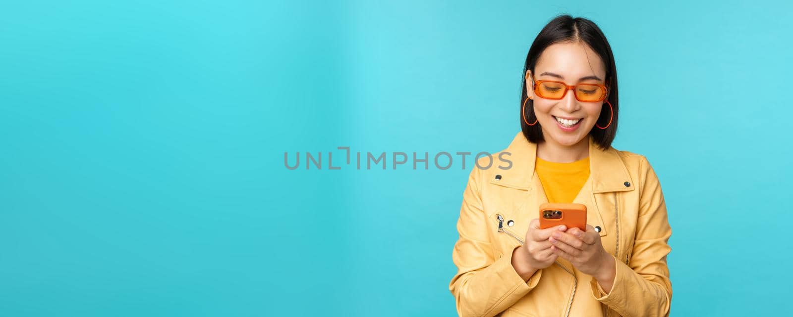 Smiling asian girl in sunglasses, using smartphone app, holding mobile phone, standing over blue background. Copy space