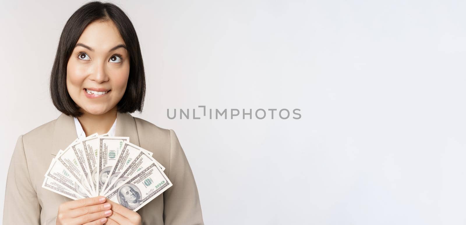 Image of asian corporate woman, happy businesswoman showing money, cash dollars and thinking, standing in suit over white background.