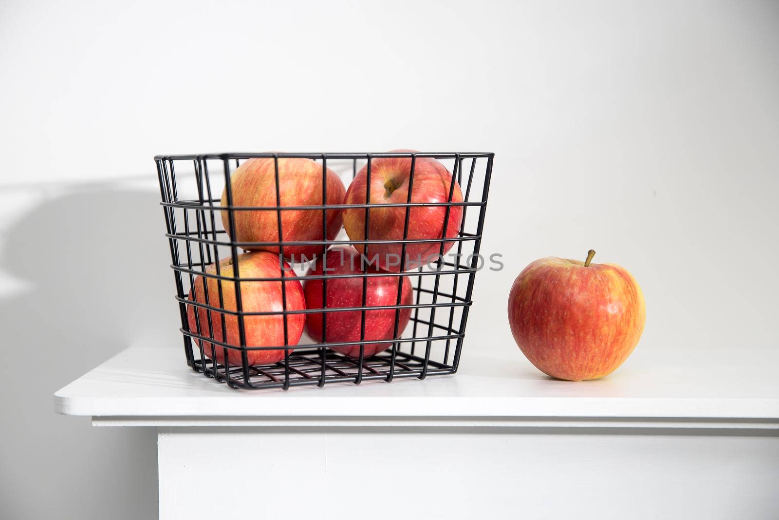Five red apples in a black metal storage basket that stands on a white mantel by elenarostunova