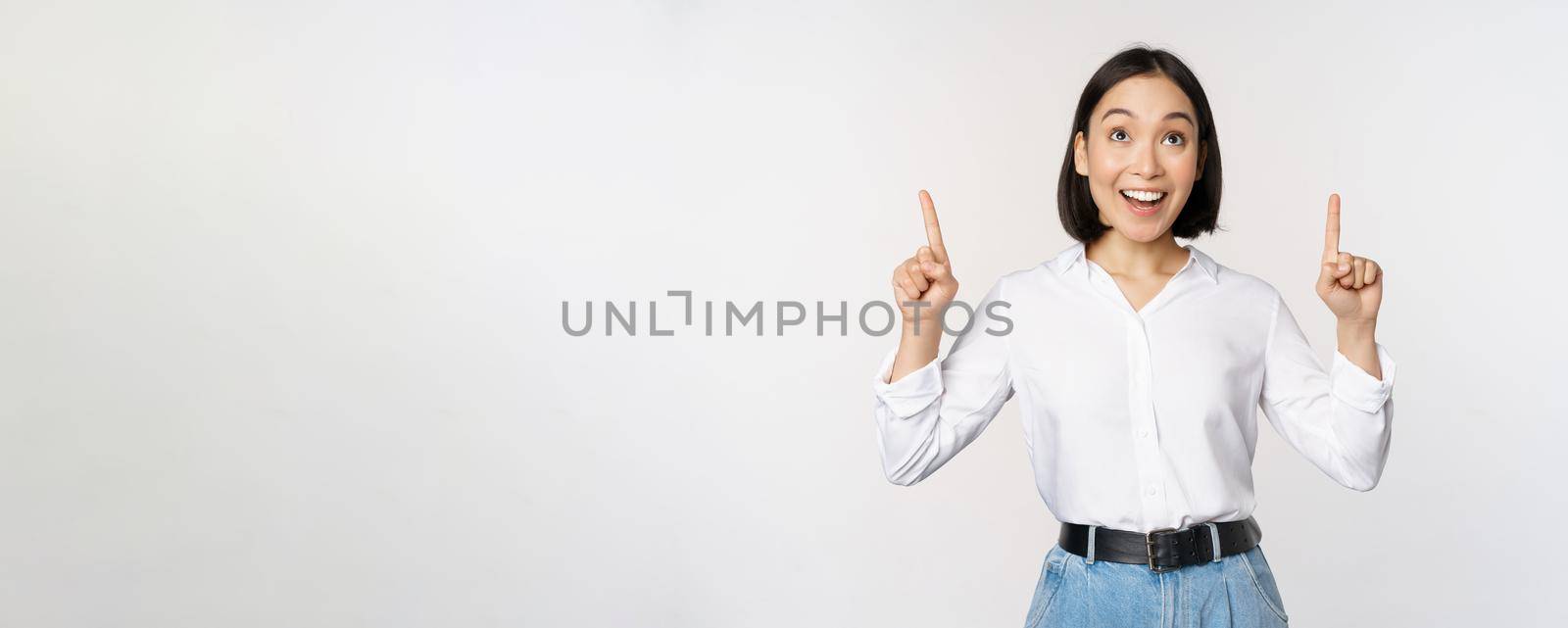 Enthusiastic asian business woman pointing, looking up with happy smiling face, showing company logo or banner, standing over white background.