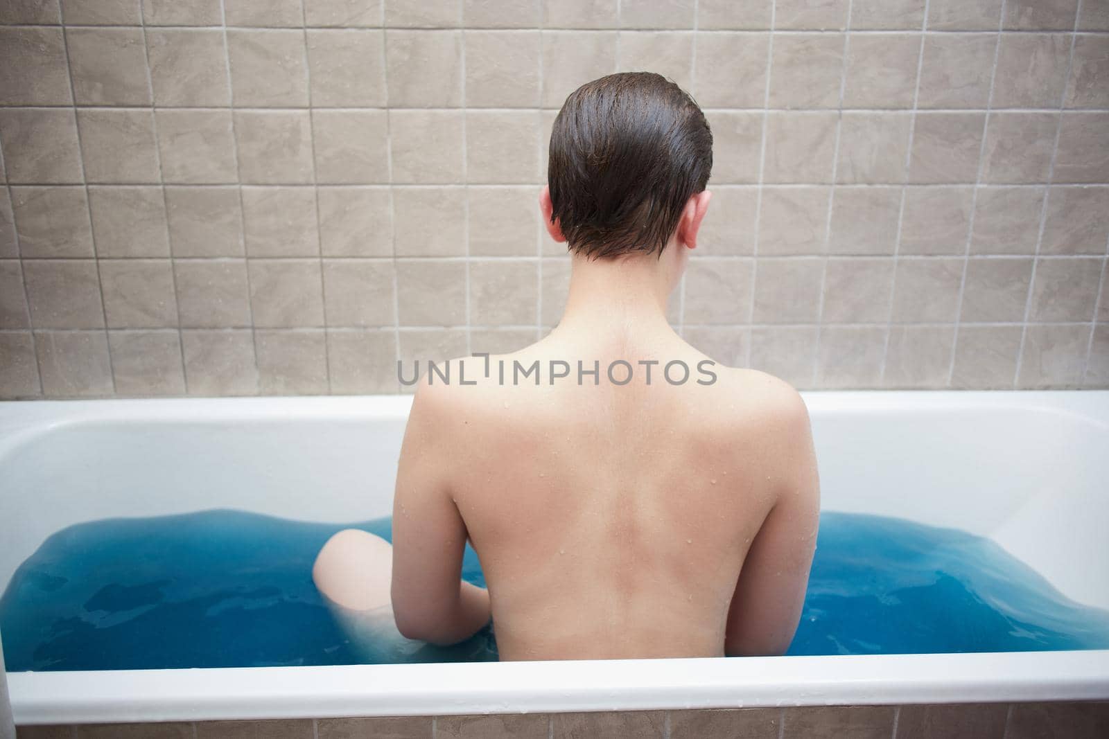Unrecognizable girl with wet, smoothly combed hair sits in blue water in the bathroom with her back against the background of beige ceramic tiles