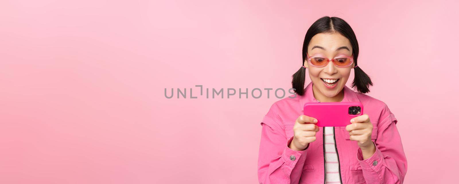 Portrait of happy asian girl playing on smartphone, watching videos on mobile phone app, standing over pink background.