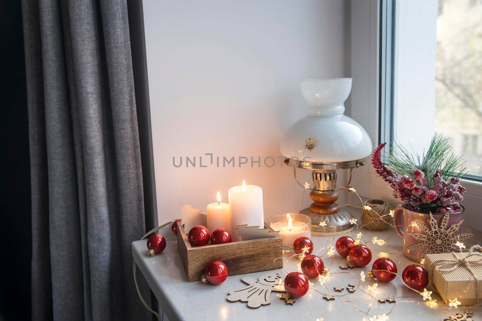 An artificial spruce branch, decorated with ivy leaves, apples and hawthorn berries. Lighted candles in wooden box, red glass balls, winter composition decorates interior on eve of Christmas by elenarostunova