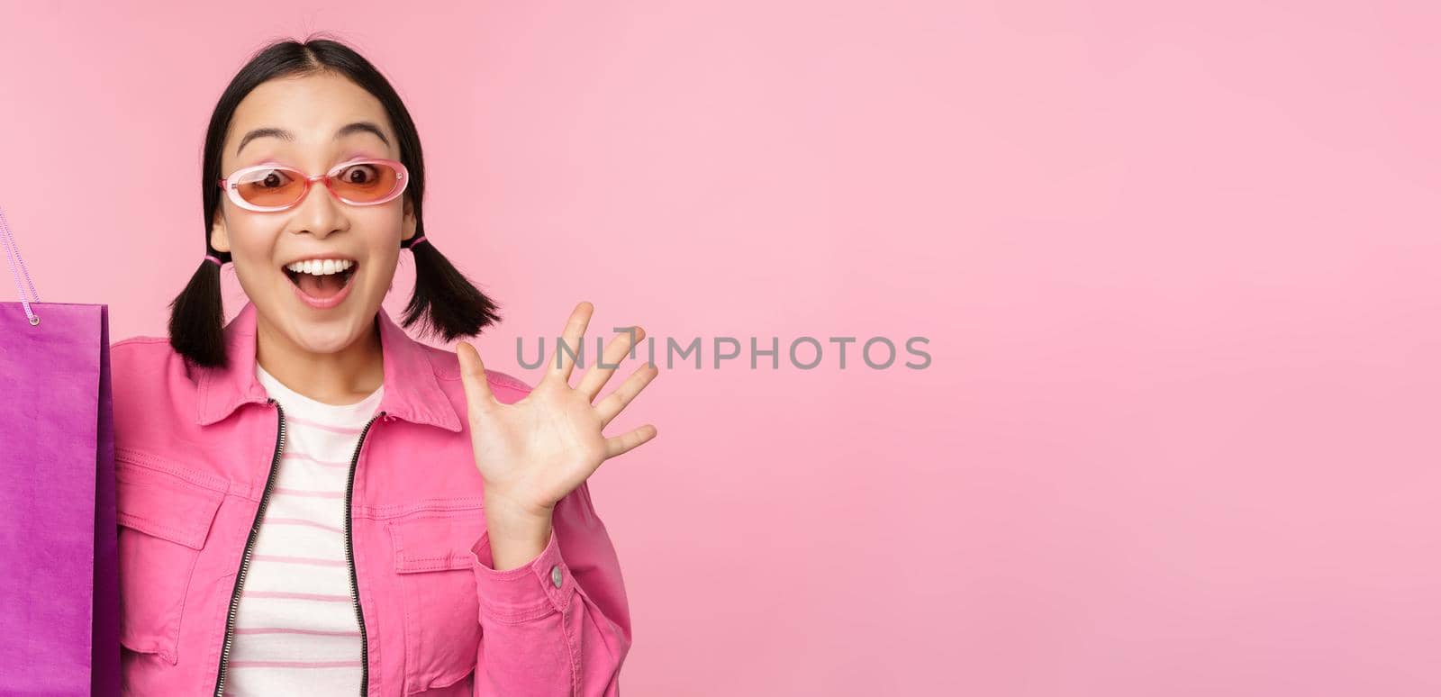 Shopping. Stylish asian girl in sunglasses, showing bag from shop and smiling, recommending sale promo in store, standing over pink background.