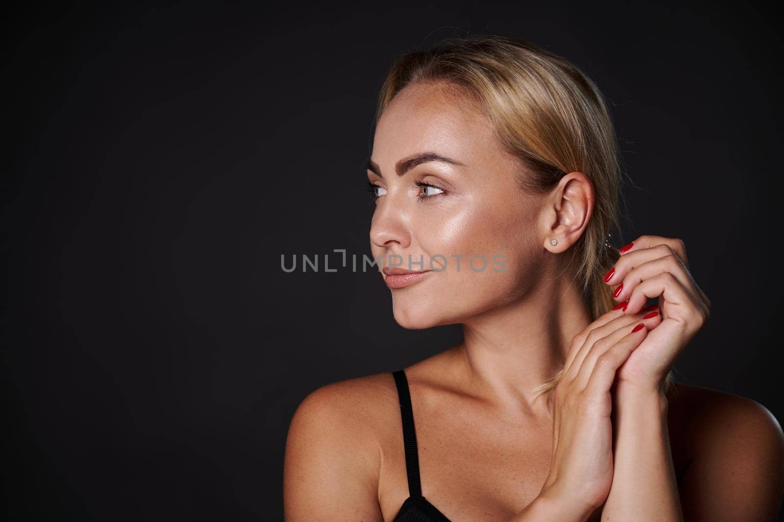 Attractive stunning well-groomed young Caucasian pretty woman with blond hair, radiant healthy tanned skin and well-groomed manicure looks aside posing on a black background with copy ad space for ads