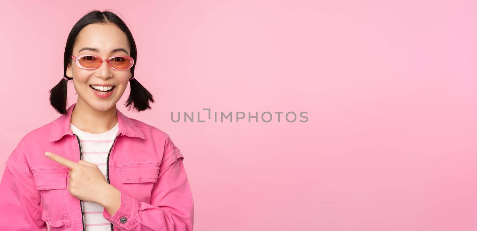Portrait of smiling asian girl in stylish outfit, sunglasses, pointing finger left, showing advertisement, banner, standing over pink background by Benzoix