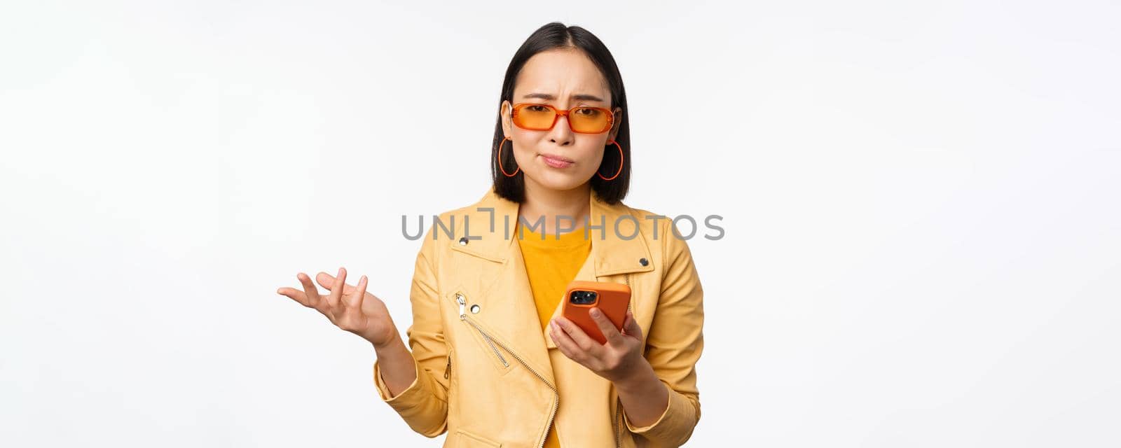 Portrait of confused asian girl in sunglasses, holding smartphone, using mobile phone and looking puzzled, standing clueless over white background by Benzoix