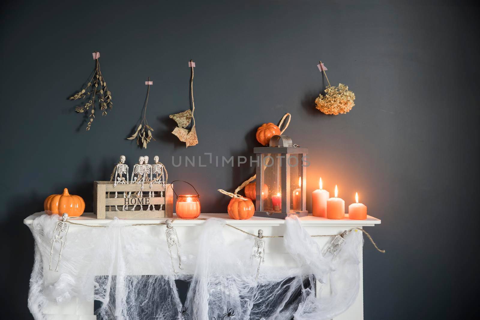 Halloween home decoration. Plastic toy skeletons in a wooden box on a fireplace against a dark blue wall. A garland of skeletons. Cobweb on the dresser. Orange candles and lantern.