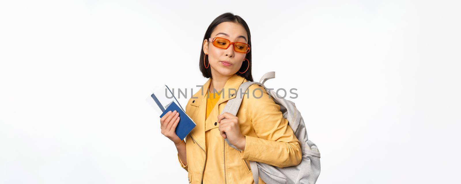Stylish asian woman in sunglasses going abroad with backpack, tourist holding passport and flight boarding tickets, standing over white background. Copy space